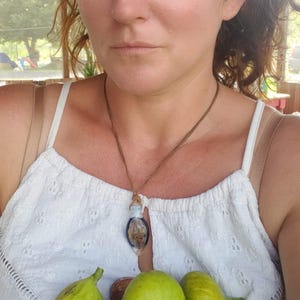 May include: A white sleeveless top with eyelet embroidery and a brown cord necklace featuring a small, oval-shaped pendant with a dark blue border. The garment is held against a collection of fresh, whole green and dark purple figs.