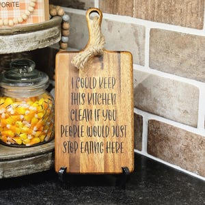 May include: Wooden cutting board with a handle and rope detail. The board has the text "I COULD KEEP THIS KITCHEN CLEAN IF YOU PEOPLE WOULD JUST STOP EATING HERE" etched into the wood. The board is displayed on a black stand.