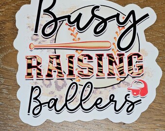 May include: A white sticker with the words "Busy Raising Ballers" in black script and bold block letters. A baseball bat and ball graphic are in the center. A red baseball helmet is in the lower right corner.