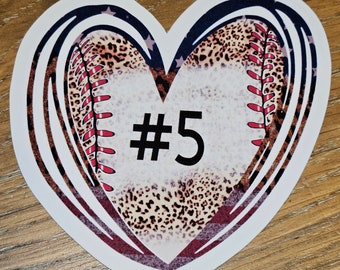 May include: Heart-shaped sticker featuring a baseball design with a leopard print center and red stitching. The number "#5" is prominently displayed in black. The sticker has a patriotic theme with blue and white stripes and stars.