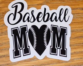 May include: A white sticker with the words "Baseball Mom" in a script font. The word "MOM" is in bold, block letters. A black baseball-shaped heart is in the center. The sticker is on a wooden surface.