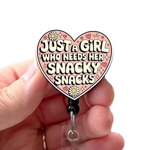 May include: A heart-shaped badge reel with the text "JUST A GIRL WHO NEEDS HER SNACKY SNACKS" in a retro font. The badge reel is pink, white, and black, with floral and heart accents. It has a retractable cord and a belt clip.
