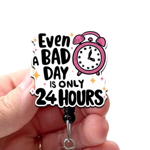 May include: A white badge reel with the text "Even a bad day is only 24 hours" and a pink alarm clock graphic. The badge reel has a black cord and a clear plastic holder for an ID badge.