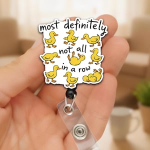 May include: A white badge reel with a cartoon design of yellow ducks and the text "most definitely not all in a row". The badge reel has a clear plastic strap and a black retractable cord.