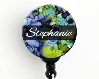 May include: A black and clear retractable badge holder with a round, colorful, abstract design and the name "Stephanie" printed in white on a black stripe.