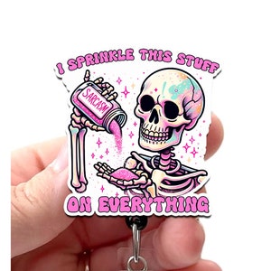 May include: A white and pink skeleton with a pink bottle labeled "Sarcasm" is sprinkling pink glitter from the bottle onto its hand. The text "I sprinkle this stuff on everything" is written in pink around the skeleton. The skeleton is on a white background. The image is a badge holder.