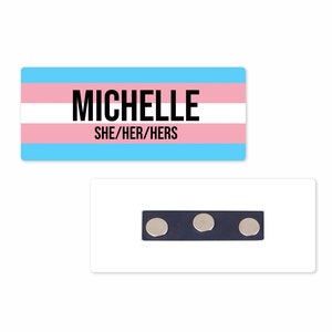 May include: A blue, pink, and white name tag with the text "MICHELLE SHE/HER/HERS" printed in black. The name tag has three silver magnets on the back.