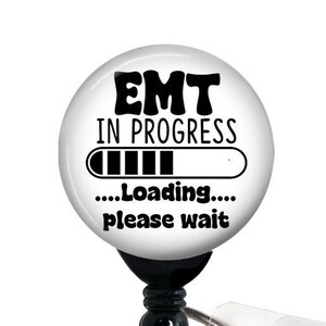 May include: White round badge holder with a black retractable clip. The badge features a black and white graphic that reads "EMT in progress...Loading...please wait".