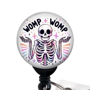 May include: A white round badge holder with a black skeleton with rainbow colored bones and the words "WOMP WOMP" in black text. The badge holder is attached to a clear plastic retractable badge reel.