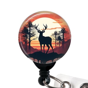 May include: A black retractable badge reel with a round, colorful button featuring a silhouette of a deer standing in a forest at sunset.