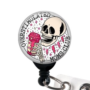 May include: A round badge reel with a skull graphic holding a pink drink, with the text "OVERSTIMULATED MOMS CLUB". The badge reel is black with a clear plastic strap and a metal snap.