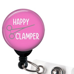 May include: A pink badge reel with the words "HAPPY CLAMPER" in white, along with a gray illustration of surgical scissors. The reel has a black backing, a silver clip, and a clear plastic strap.