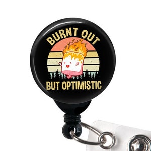 May include: Black and white badge reel with a black button and a clear plastic strap. The badge holder features a retro-style graphic of a burning marshmallow with the text "Burnt Out But Optimistic".
