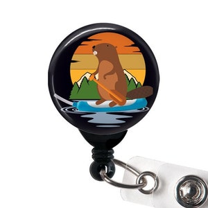 May include: A black retractable badge holder with a round, colorful design featuring a cartoon beaver paddling on a surfboard in front of a sunset. The beaver is brown and has a white belly. The surfboard is blue and has a yellow paddle. The sunset is orange and yellow with a dark blue sky. The badge holder has a clear plastic strap.