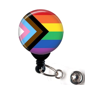 May include: A black retractable badge reel with a round, colorful button featuring a rainbow pride flag with a black and brown chevron.