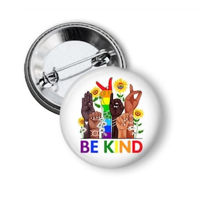 May include: A white button with a rainbow-colored design featuring hands in different gestures and the text "BE KIND". The design also includes sunflowers and daisies.
