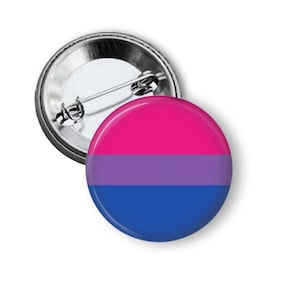 May include: A round button with a pin back featuring the bisexual pride flag. The flag has three horizontal stripes: pink, purple, and blue.