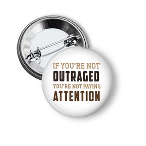 May include: A white button with the text "IF YOU'RE NOT OUTRAGED YOU'RE NOT PAYING ATTENTION" in brown lettering. The button has a silver metal backing with a pin.