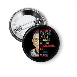 May include: A round, black button with a silver pin back. The button features a colorful portrait of a woman and the text "WOMEN BELONG IN ALL PLACES WHERE DECISIONS ARE BEING MADE."