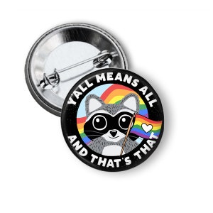 May include: A black button with a raccoon holding a rainbow pride flag. The text around the raccoon says "Y'all Means All And That's That".