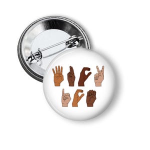 May include: A white button pin with a metal backing. The pin features a design of hands spelling out "ASL LOVE" in American Sign Language. The hands are depicted in various skin tones against a white background.