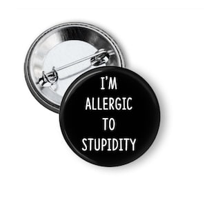 May include: A black button pin with the text "I'M ALLERGIC TO STUPIDITY" in white. The pin has a metal backing with a safety pin style clasp.