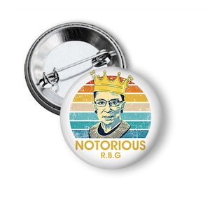 May include: A white button with a retro-style image of Ruth Bader Ginsburg wearing a crown. The image is set against a colorful sunset background. The text "NOTORIOUS R.B.G." is printed below the image.