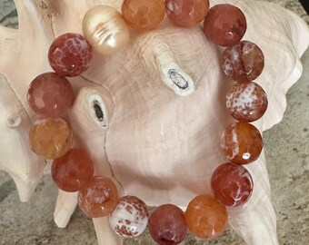 May include: A beaded bracelet featuring faceted, round beads in shades of orange, red, and white. The bracelet includes a single, oval-shaped pearl. The jewelry is displayed on a light-colored seashell.