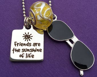 May include: A silver necklace with a charm that says "Friends are the sunshine of life" and a sun graphic. The necklace also features a yellow and silver bead and a sunglasses charm with black lenses.