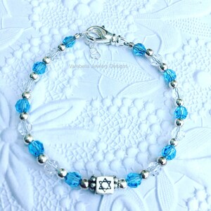 May include: A silver bracelet with blue and clear crystal beads and a silver charm with a Star of David. The bracelet is on a white background. Valebella Jewelry Designs.