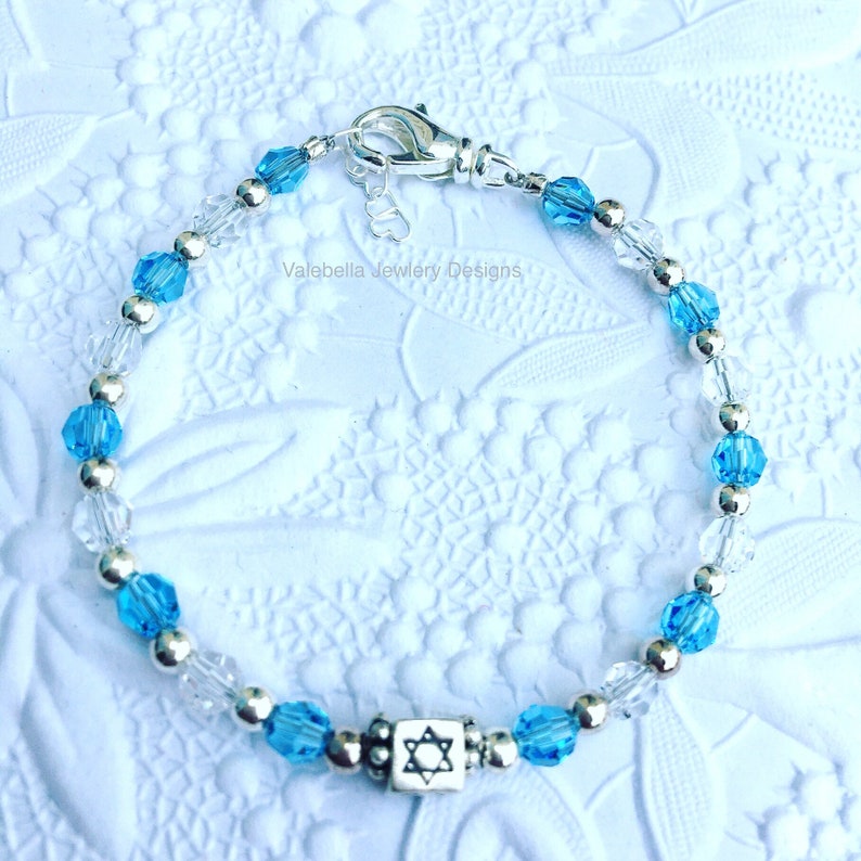 May include: A silver bracelet with blue and clear crystal beads and a silver charm with a Star of David. The bracelet is on a white background. Valebella Jewelry Designs.