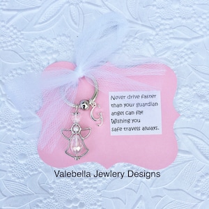 以下が含まれることがあります： 小さなガラス瓶に2つの小さなハートが入った、銀色の天使のキーホルダー。キーホルダーは、ピンク色のカードに付けられており、カードには「あなたの守護天使が飛ぶよりも速く運転しないでください！安全な旅を願っています。」というテキストと「Valebella Jewelry Designs」というテキストが書かれています。