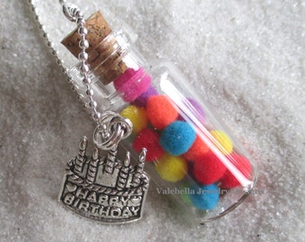 May include: A clear glass vial necklace filled with colorful pom-poms, sealed with a cork, and a silver-tone charm that says "HAPPY BIRTHDAY" in a cake design. The necklace chain is a silver ball chain.
