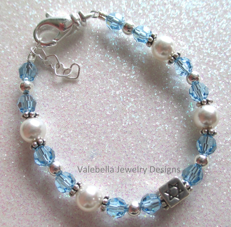 May include: A silver bracelet with blue crystal beads and white pearls. The bracelet has a silver charm with a Star of David design. The bracelet is on a sparkly background.