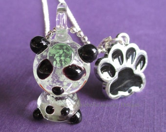 May include: A silver chain necklace with two glass charms. One charm is a panda bear with black and white spots and a green and white swirl inside. The other charm is a black paw print with a silver outline.