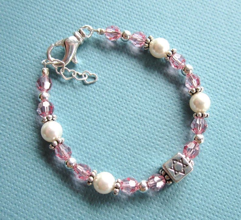 May include: A pink and white beaded bracelet with a silver Star of David charm. The bracelet is made with small pink glass beads, white pearls, and silver beads.