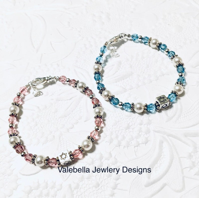 May include: Two beaded bracelets with silver clasps and charms. One bracelet has pink and white beads, the other has blue and white beads. Both charms are silver and feature a Star of David.