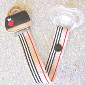 May include: A black and white striped pacifier clip with a red stripe. The clip features a felt handbag with a red heart and white accents.