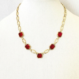 May include: A gold-tone chain necklace featuring five rectangular, faceted red glass stones set in metal frames. The necklace is constructed with alternating oval-shaped links and is displayed on a neutral-colored, textured fabric bust form.