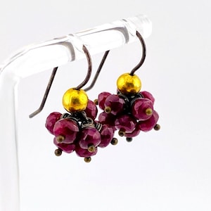 May include: A pair of dangle earrings featuring clusters of small, faceted, deep magenta-colored beads. Each earring is topped with a single smooth, round, metallic gold bead and hangs from a dark, oxidized metal hook wire for a secure fit.