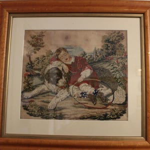 May include: A framed needlepoint embroidery depicting a young child sleeping with a dog. The child is wearing a red dress and the dog is white with black spots. The scene is set in a grassy field with trees and a river in the background.