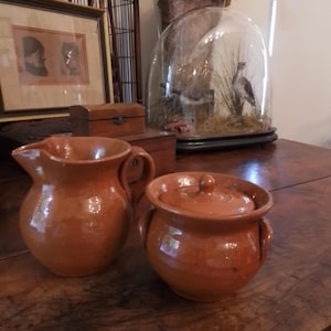 May include: A terracotta pitcher and a matching lidded pot with two side handles. Both pieces feature a smooth, warm brown clay finish with a subtle sheen. The pitcher has a curved spout and a single handle, while the pot includes a round lid.