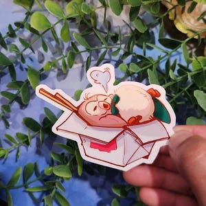 May include: A cartoon sticker of a pink and white penguin wearing a green bow tie and sleeping in a white takeout container with chopsticks sticking out. The container has a red and white label.