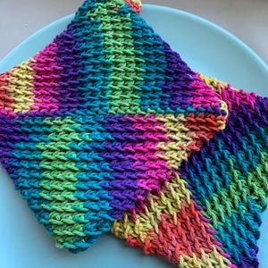 May include: Two colorful crocheted dishcloths in a rainbow pattern. The dishcloths are made with a textured stitch and are folded on top of each other. The dishcloths are on a light blue plate.