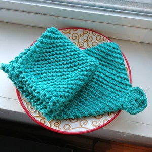 May include: Two teal blue knitted dishcloths on a white plate with a red and gold rim. The dishcloths are made with a simple stitch pattern.