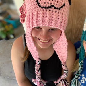 May include: A pink crocheted hat with a smiling face and black eyes. The hat has long pink and white braided ties.