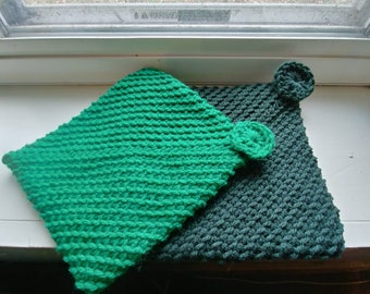 May include: Two handmade, square potholders. One is bright green, the other is a darker teal green. Each has a small, circular decorative element attached to one corner. The potholders are made of a thick, knitted material.