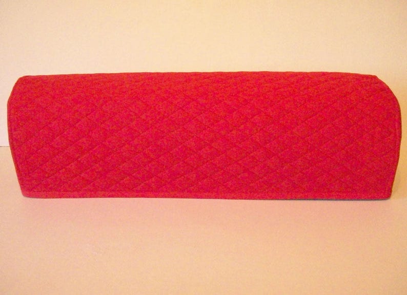 May include: A quilted, red fabric cover with a diamond pattern. The cover has a rectangular shape with rounded edges. The item is likely a cover for a kitchen appliance, such as a toaster oven or microwave.
