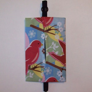 May include: A fabric tissue holder with a blue, green, and yellow background featuring red birds and white flowers. The holder has a black strap for hanging.