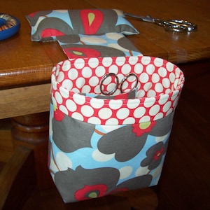 May include: A gray and blue fabric hanging organizer with white polka dots and red flowers. The organizer has a pocket on top and a hanging pouch underneath. The pouch contains two pairs of silver scissors.